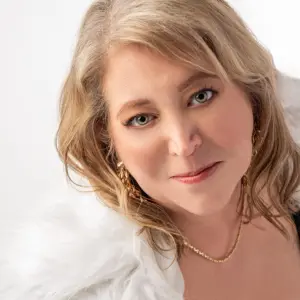 A smiling woman over 40 with blonde hair, natural glam makeup, gold earrings, and a white fur wrap in a studio portrait.
