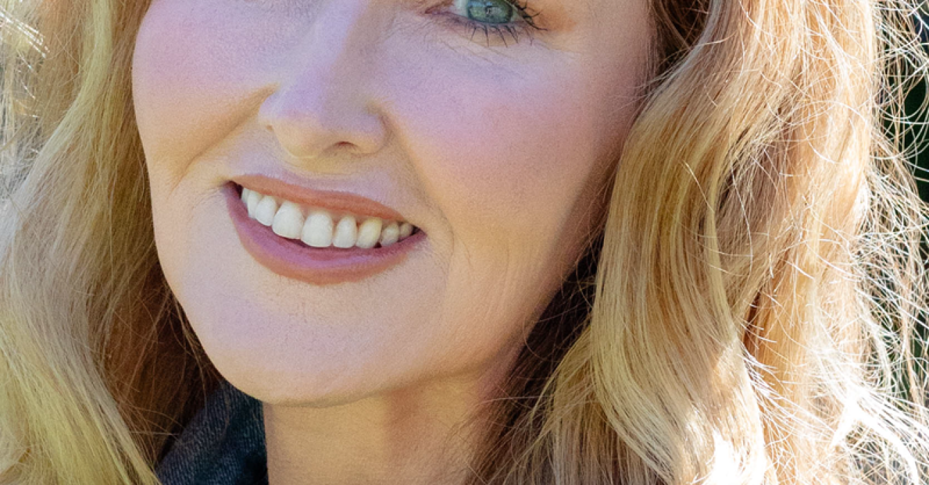 A smiling woman over 50 with radiant satin skin, natural pro-aging makeup, and blonde wavy hair, showcasing a healthy glow.